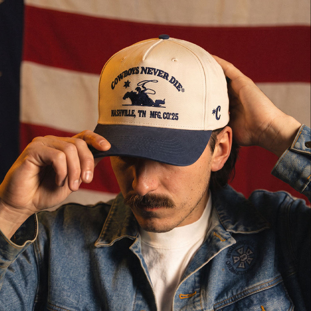 A man wearing a cream and navy blue baseball cap with 'Cowboys Never Die' and 'Austin, TX NFL' printed on the front, holding the brim with one hand and adjusting the side strap with the other, set against a blurred American flag background.
