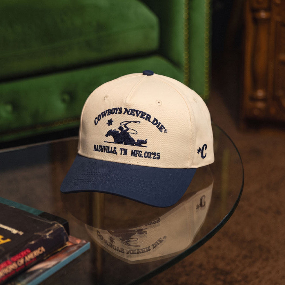 A baseball cap resting on a glass table in a living room setting, featuring a cream crown with navy blue text and graphic reading 'COWBOYS NEVER DIE' and 'BAILORRALL, TX MFG. CO.