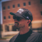 A man wearing a black baseball cap with white text that reads 'GODS BY DOGS 2025' and a small white graphic on the side, along with black sunglasses and a dark gray shirt, photographed outdoors with a blurred brick building in the background.