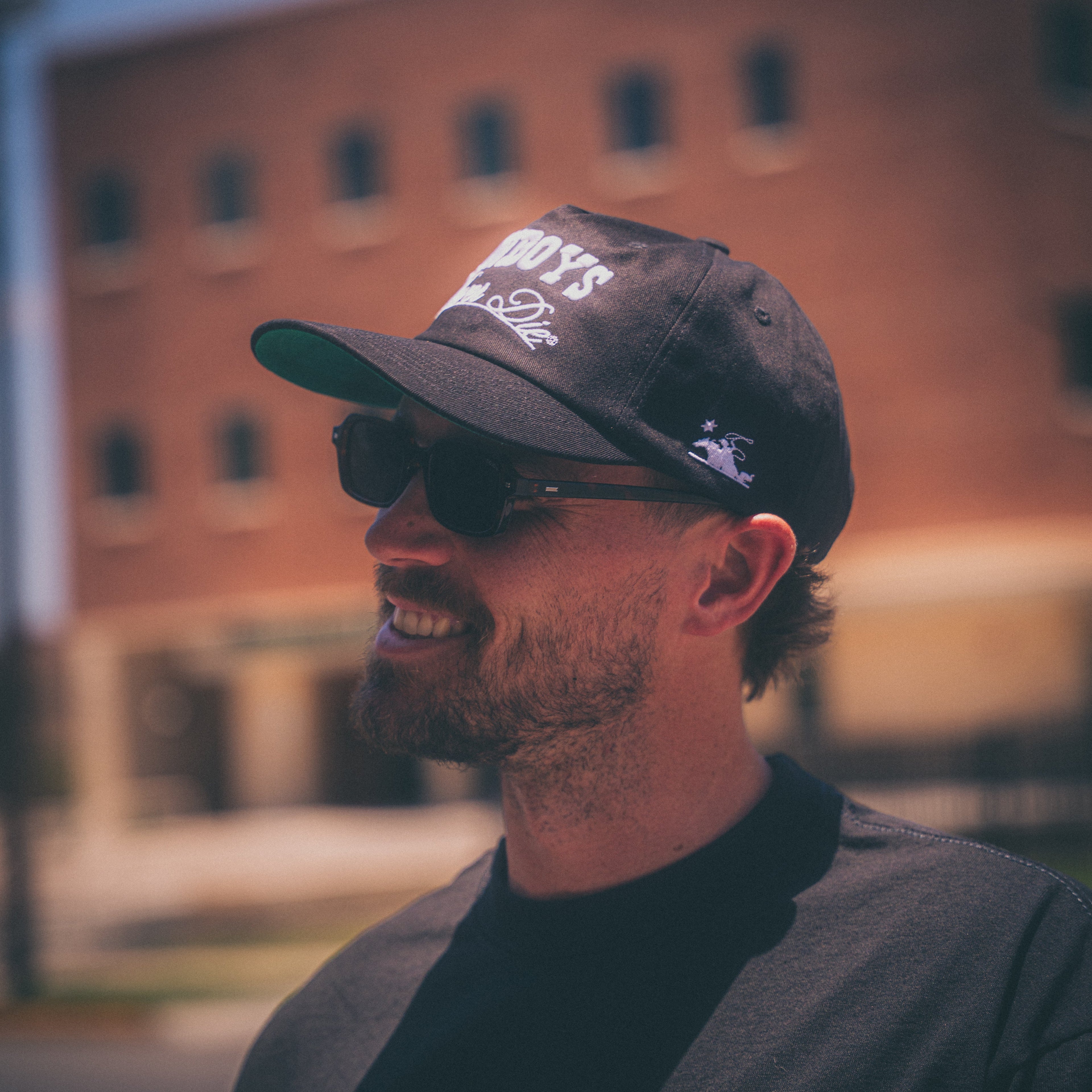 A man wearing a black baseball cap with white text that reads 'GODS BY DOGS 2025' and a small white graphic on the side, along with black sunglasses and a dark gray shirt, photographed outdoors with a blurred brick building in the background.