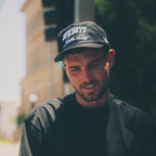 A man wearing a black t-shirt and a black baseball cap with 'COWBOYS' written on it, standing on an outdoor street with blurred buildings in the background.